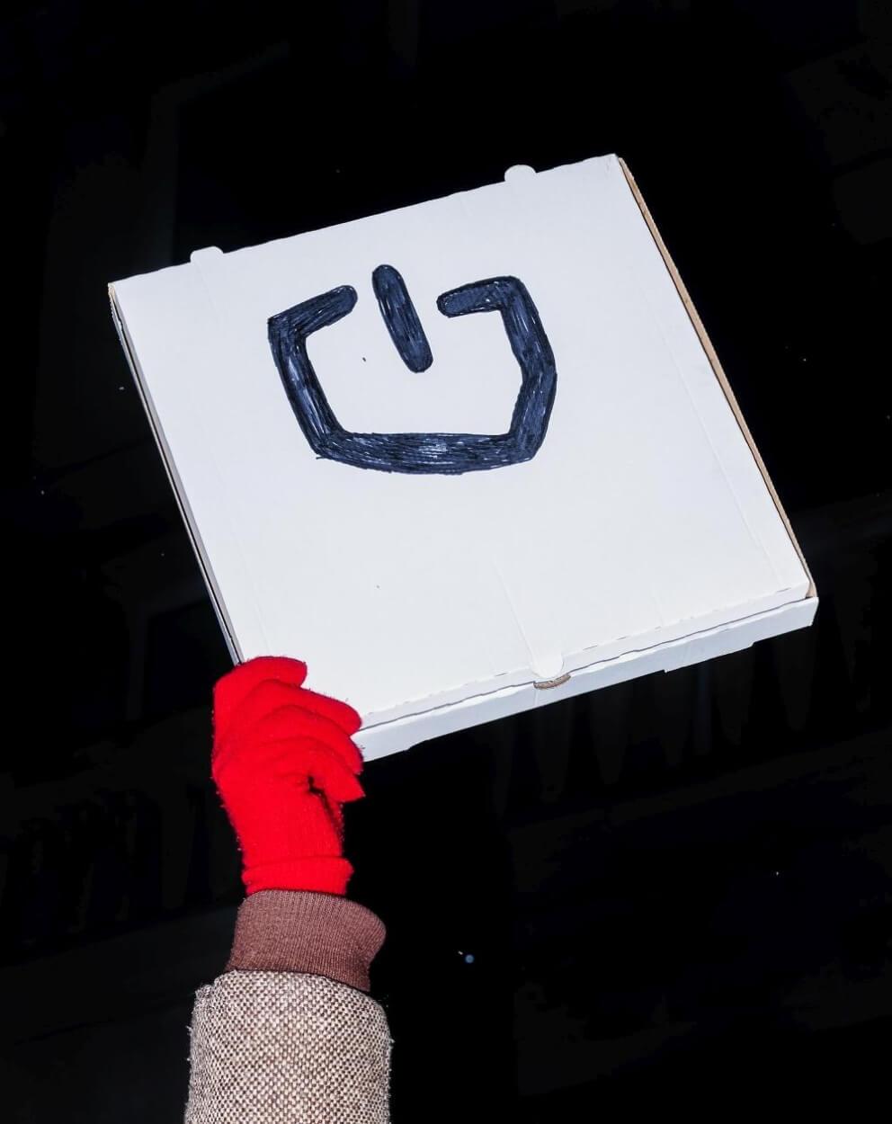 Hand holding a protest sign drawn on a pizza box; the sign depicts a computer power symbol in the shape of Poland